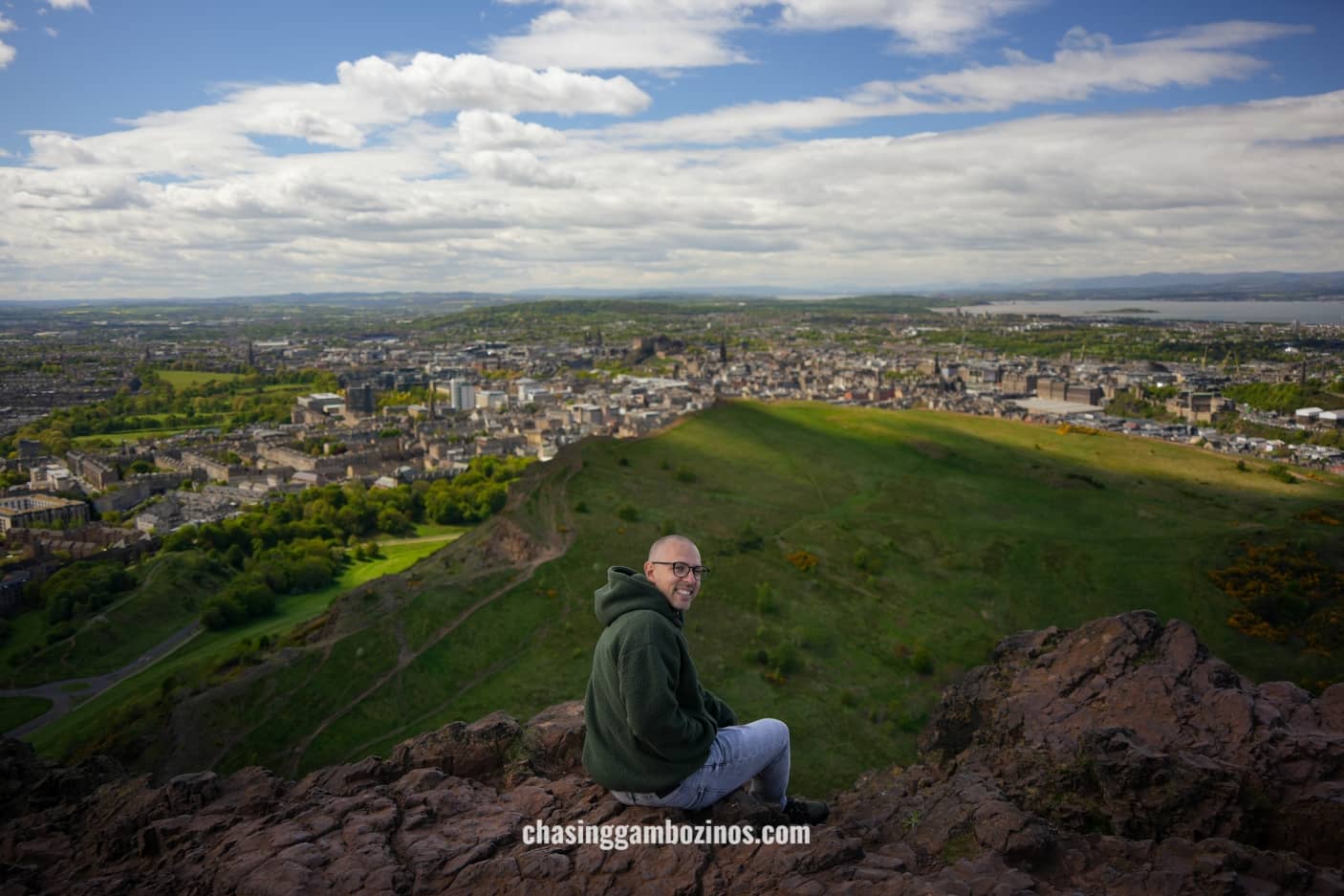 Homem a sorrir no topo do Arthur’s Seat, com vista sobre a cidade em fundo.