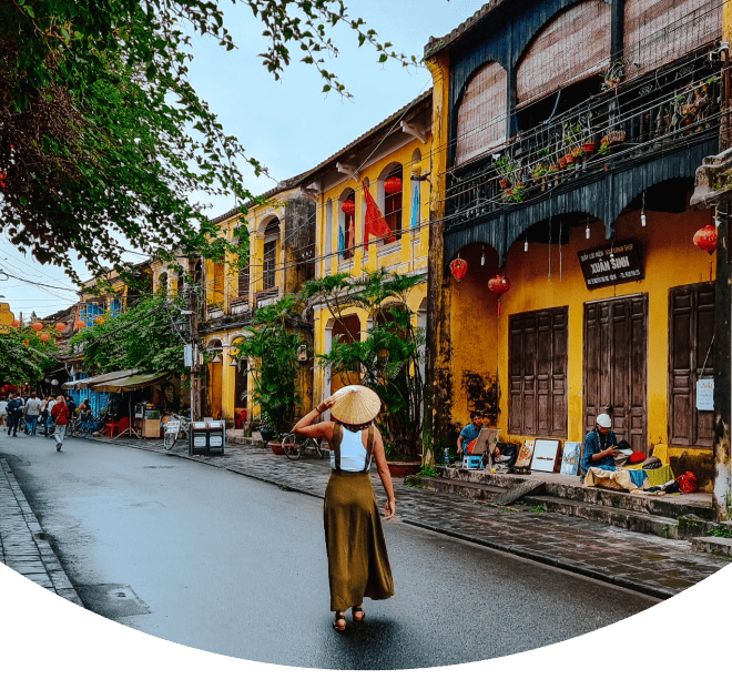 Woman wearing a traditional hat walking down a street of yellow houses in Hoi An, Vietnam. A must visit city in any Vietnam itinerary.